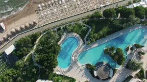 Een foto vanuit de lucht genomen. Je zit een vakantieparadijs, met zwembaden, palmbomen en een strand met strandstoelen. In een hoek van het beeld zie je ook nog een stukje zee.