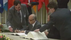 Een foto van twee mannen die achter een tafel zitten. Op die tafel ligt een heel dik open boek. Een man zet zijn handtekening op een document. Twee mannen die erbij staan kijken aandachtig mee. Op de achtergrond zie je vlaggen van landen.