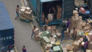 Een foto van een groep mannen en tientallen dozen. Enkele mannen zijn een vrachtwagen aan het inladen of uitladen. De dozen die buiten staan staan kriskras door elkaar. 
