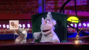 Een foto van de pop van Nico de Neushoorn met een bril. Hij zit op een groene stoel achter een bureau met een boek erop. Naast de neushoorn is de pop Stella Schaap te zien. Stella heeft een bord vast met daarop een getekende mannetjesleeuw. 