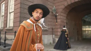 Een foto van presentator Nizar in een ouderwets kostuum. Hij draagt een lichtbruin pak met knopen, een lichtbruine mantel van dezelfde kleur en een zwarte hoed met witte veer. Daarachter loopt iemand in ouderwets kostuum naar de poort van een oud gebouw.