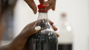 Een foto van twee handen die een fles met cola openmaken. Eén hand heeft de fles vast en met de andere hand wordt de rode dop van de fles gedraaid. Als het normale cola is met veel bubbels wat CO 2 is erin, zal de cola uit de fles gaan spuiten.
