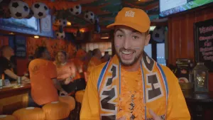 Een foto van de presentator met oranje kleren aan. Hij staat in een café en kijkt in de camera. Het café is helemaal versierd voor het Oranjegevoel. Alle mensen in het café hebben oranje kleren aan en er hangen voetballen aan het plafond.