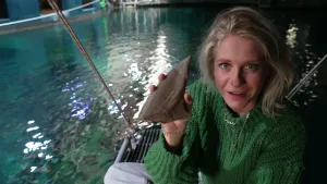 Een foto van Janouk die naast een groot aquarium, een basin, in de dierentuin in Arnhem zit. In haar rechterhand houdt ze een bruingrijze rugvin van een haai omhoog naar de camera. Janouk kijkt in de camera. Ze draagt een groene trui en lichtblauwe broek.