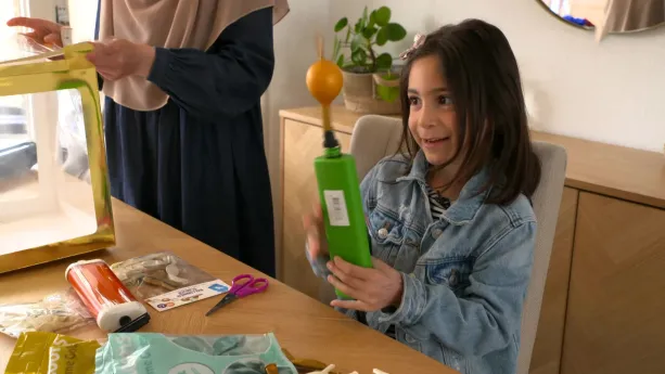 Een foto van een meisje van ongeveer zeven jaar oud. Ze zit aan een tafel en heeft een apparaat in haar handen. Dat apparaat blaast een goudkleurige ballon op. Naast haar staat een vrouw met Islamitische kleren aan.