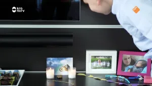 Een foto van een jongetje dat voorovergebogen rechts in beeld staat. Hij kijkt naar een kastje waarop twee brandende kaarsen staan. Daarvoor ligt een kettinkje met een kruis eraan. Op het kastje staan ook enkele foto's van kinderen.