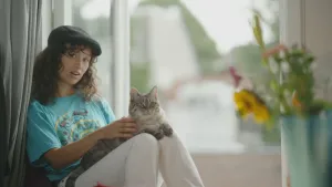 Een foto van de presentator. Ze zit op de grond en leunt tegen een muur. Op haar benen zit een kat. Ze houdt de kat voorzichtig vast en kijkt in de camera.