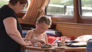 Een foto van een vrouw en een jong meisje binnen in een boot. Het meisje zit aan tafel. Daarop liggen allerlei spullen, waaronder een boterham. De vrouw kijkt naar wat het meisje aan het doen is.