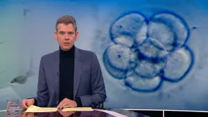 Een foto van de presentator die achter een bureau zit. Hij vertelt iets in de camera. Op de achtergrond zie je een grote foto van een embryo. Dat is een groepje cellen waarmee een mensenleven begint.