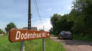 Een foto van een landschap met op de voorgrond een bruin wegwijzerbord. In witte letters staat daarop de tekst "Dodendraad WO 1" en een pijl naar rechts. Je ziet een auto op een weg die kant op rijden. 