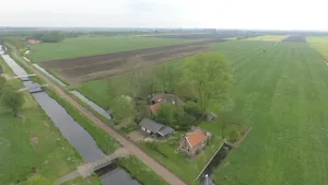 Een luchtfoto van weilanden met voorop een aantal woningen die langs een weg en water kanaal staan.