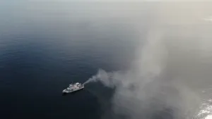 Een foto van een schip in een verder lege zee. De foto is vanuit de lucht genomen. Het schip blaast rook de lucht in. Dat geeft een lichtgrijze wolk boven het water.