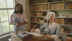 Een foto van presentator Tirsa. Ze staat in een kamer in een oud huis en kijkt naar de camera. Naast haar staat een oude schrijftafel met daarachter een grote pop die een vrouw moet voorstellen. Die vrouw is een schrijver van lang geleden.