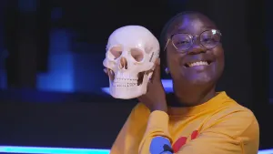 Een foto van presentator Tirsa die in de camera kijkt. Ze houdt een plastic schedel naast haar hoofd. Tirsa laat haar tanden zien, en van die schedel zie je natuurlijk ook de tanden. 