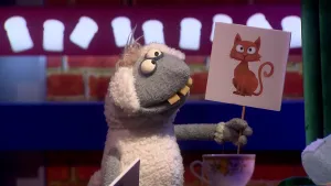 Een foto van pop Stella Schaap. De pop heeft een bord met een getekende bruine kat erop vast en kijkt naar het bord. De pop heeft een witte vacht op haar rug en oren, een grijze vacht bij kop en poten, ronde ogen en vooruitstekende, gele tanden.