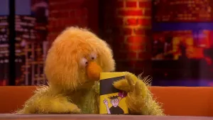 Een foto van een pop van een presentator met een gele vacht en grote gele neus die een boek vastheeft. Het boek, geschreven door pop Lucas, is geel en zwart met een tekening van een mens erop die de vinger in de lucht steekt en een uitroepteken erboven. 