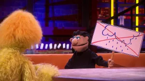 Een foto van twee poppen, een presentator met een gele vacht links, en meneer Korzel rechts aan een tafel. Meneer Korzel zit op een bruine bank. Hij protesteert tegen het weer met een bord, waarop een wolk met regen en een rood kruis erdoor staan.