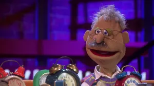 Een foto van pop Tom Roelofs. Tom verzamelt wekkers en daarvan staan zeven wekkers voor hem op de tafel. Het zijn ouderwetse wekkers met wijzers, twee belletjes op de bovenkant en een drukknop ertussenin. Tom heeft grijs haar, een snor en een bril op.