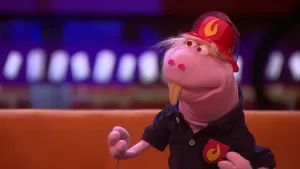 Een foto van pop Brandweerman Fokko Slang die op een bruine bank zit. Fokko is een roze slang met twee grote, uitstekende tanden. Hij heeft brandweerkleding aan: een blauwe blouse met een roodgele vlam erop en een rode helm met een roodgele vlam erop.