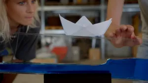 Een foto van een bootje dat met wit papier gevouwen is. Het bootje staat op een doorzichtig stuk plastic, waar onderin blauw vocht zit, op een zwarte standaard. Janouk staat gebogen erachter en Hans, waarvan de hand zichtbaar is, staat er ook achter.