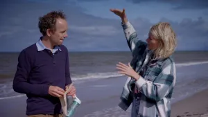 Een foto van Janouk en een wetenschapper op het strand van Texel. De wetenschapper heeft een doorzichtige fles, een vel papier dat erin zat en een drinkfles met blauwgroene glitters vast. Janouk houdt haar rechterhand hoog geheven boven de linkerkand.