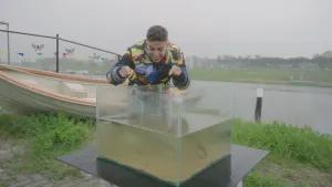 Een foto van presentator Nizar. Hij staat buiten achter een glazen bak met water. Hij wijst met beide handen naar die bak en kijkt er in. Er lijken vissen in te zwemmen. Op de achtergrond zie je een rivier.