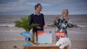 Een foto van Janouk en een man op het strand achter een tafel met een bord waar bovenaan rood en eronder blauw vocht zit, links een blauw bord "Bonaire", een palmboom en een fles met bericht, rechts een rood bord "Noordpool" en een ijsbeerknuffel. 