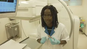 Een foto van presentator Tirsa in een behandelkamer in een ziekenhuis. Ze heeft een witte jas aan met blauwe handschoenen. In een hand houdt ze een speciaal spuitje vast. Achter en naast haar staat een groot wit apparaat met een computerscherm erop.