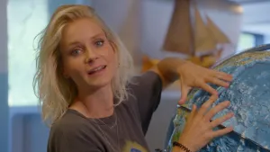 Een foto van Janouk die naast een gedeeltelijk zichtbare wereldbol staat. Janouk wijst met handen met gestrekte vingers naar de oceaan. Het is een wereldbol waar uitstekende stukjes op de plek van landen en andere opvallende delen van de wereld zitten. 