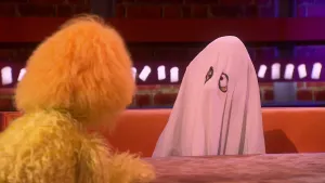 Een foto van een pop, Joop Spook, die onder een wit laken met gaten voor de ogen zit. Hij zit op een bruine bank achter een tafel en kijkt naar een geel behaarde pop waarvan links de rug te zien is. Joop is al 30 jaar spook en wil graag iets anders doen.