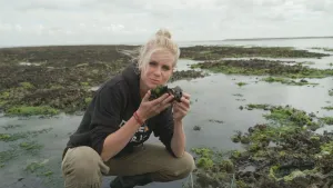 Een foto van presentator Janouk. Ze zit gehurkt in een gebied vlakbij de zee. Ze houdt een mossel in haar handen en kijkt in de camera. Op de achtergrond zie je water maar ook drooggevallen stukken land.