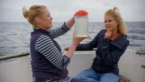 Een foto van presentator Janouk en Katja Peijnenburg, een zeebioloog die veel over de zee weet. Ze zitten aan de rand van een witte boot. Katja houdt een witte plastic pot omhoog waar water inzit, maar ook veel zeer kleine diertjes en plantjes, plankton.