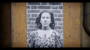 Een zwart-wit foto van Truus van Lier, een Joods-Nederlandse vrouw die tijdens de Tweede Wereldoorlog in het verzet zat. Ze staat voor een muur van bakstenen en heeft een donkere jurk aan met witte bloemetjes erop. De foto hangt op een houten achtergrond.