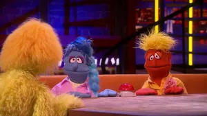 Een foto van twee poppen, Truus en Wim met voor hen een tafel met een rode drukknop erop. De drukknop staat er, omdat ze overal een quiz van maken. Truus heeft een blauwe huid en haar in een staart, Wim heeft een rode huid, scheve ogen en geel haar. 