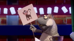 Een foto van pop Stella Schaap. De pop heeft een bord met een getekende bruine aap erop vast en kijkt naar het bord. De pop heeft een witte vacht op haar rug en oren, een grijze vacht bij kop en poten, ronde ogen en vooruitstekende, gele tanden.