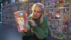 Een foto van presentator Janouk die op een tafel leunt met een doosje in haar hand. Op het doosje zijn afbeeldingen van bloemen en staat het nummer 1089 op. Op de achtergrond zijn er veel kasten met nog veel meer doosjes te zien.