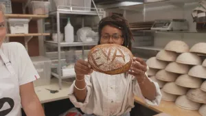 Een foto van presentator Eva die in de werkruimte staat van een bakker en een vers gebakken brood laat zien waar haar naam op staat.