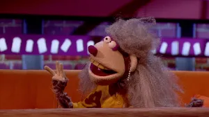 Een foto van een pop met de naam Fenna van Dale. De pop heeft  bruin haar met kleine krullen, oorbellen met zes witte bolletjes en een bruingeel vest. De neus en wenkbrauwen om de ronde ogen zijn paars. De pop zit op een bruine bank achter een tafel.