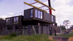 Een foto waarin een vanuit de fabriek kant en klaar huis, een prefab huis, te zien is. Een gedeelte wordt met een kraan op de geplaatste benedenverdieping gezet. Het huis heeft zwarte zijkanten en vijf gedeelten met ramen. Twee medewerkers kijken toe.