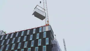 Een foto van een flat die gebouwd wordt. De flat is donkergrijs met en alle ramen zijn goog en smal. Een hijskraan staat op het dak en verplaatst een groot blok. 