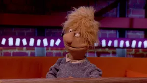 Een foto van een pop die op een bankje aan een tafel zit. Achter hem op de muur zijn witte boterham vormpjes op een paarse achtergrond, en bakstenen.