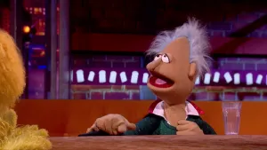 Een foto van Goochelaar Ariando, een pop die aan een tafel met  glas water erop zit. Hij heeft een lichtbruine huid, grijswitte plukjes haar en draagt een groen glittervest met rood-witte kraag. Rechts is van de achterkant een pop met gele vacht te zien.