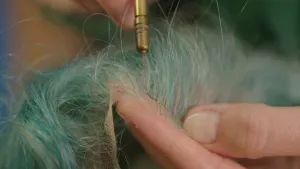 Een foto van een hand die een pruik met blauw haar vasthoudt. De andere hand houdt een stuk gereedschap tegen de pruik aan. Het is een metalen handvat met daaraan een dunne kromme naald.