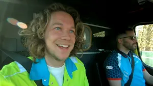 Een foto van Pascal die meerijdt in een ambulance die op weg is na een melding via noodnummer 112. Naast Pascal zit de ambulancemedewerker en bestuurder. Pascal draagt een felgroene blouse. De medewerker draagt een blauwe blouse en heeft een zonnebril op.