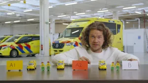 Een foto van presentator Pascal achter een tafel. Op de tafel staan kleine versies van ambulances, poppetjes en papiertjes met daarop "A1", "A2" en "B". Achter hem is een grote ruimte met een ambulance en twee auto's in dezelfde kleuren als een ambulance.