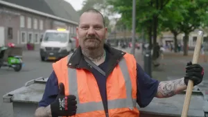 Een foto van een stratenschoonmaker, die zijn duim omhooghoudt. In zijn andere hand heeft hij een bezemsteel vast. Hij heeft een fel oranje vestje aan. Achter hem rijdt een busje voor het afval. Eromheen zijn huizen en een pleintje.