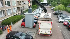 Een foto van een straat met een vuilniswagen. Een hijskraan op de wagen tilt een ondergrondse afvalbak uit de stoep. Dat is een grote grijze bak. Links op de foto staat de presentator Tirsa met een vuilnisman. Rondom staan huizen en auto's.
