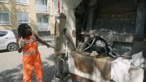 Een foto van presentator Tirsa, die bij de achterkant van een vuilniswagen staat. Ze drukt op een knop aan de zijkant en kijkt naar een voorwerp dat platgedrukt wordt in de wagen. Ze kijkt verbaasd en onder de indruk.