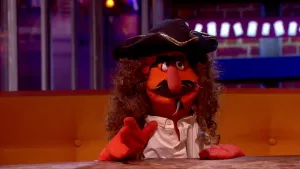 Een foto van een pop, Berend Dubbel, die tegen de camera praat. Hij heeft piratenkleding aan. Hij zit aan een tafel op een bank. 