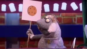 Foto van een pop, Stella Schaap. Ze houdt een bordje omhoog met een tekening van een koekje erop. Achter haar is een muur met bovenaan witte boterham vormen.